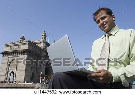 Stock Image - Portrait of a businessman using a laptop and smiling, Gateway of India, Mumbai, Maharashtra, India. Fotosearch