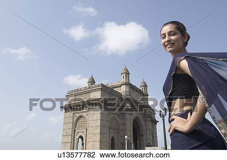Stock Image - Portrait of a young woman standing with a monument in the background, Gateway Of India, Mumbai, Maharashtra, India. Fotosearch