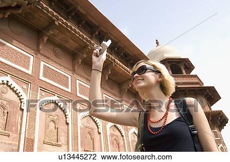 Stock Image - Low angle view of a young woman taking a picture in front of a mausoleum, Taj Mahal, Agra, Uttar Pradesh, India. Fotosearch