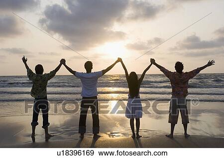 Stock Photo - Rear view of three young men and a young woman standing side by side with holding hands on the beach. Fotosearch