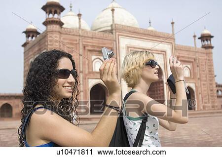 Stock Image - Side profile of two young women taking a picture in front of a mausoleum, Taj Mahal, Agra, Uttar Pradesh, India. Fotosearch
