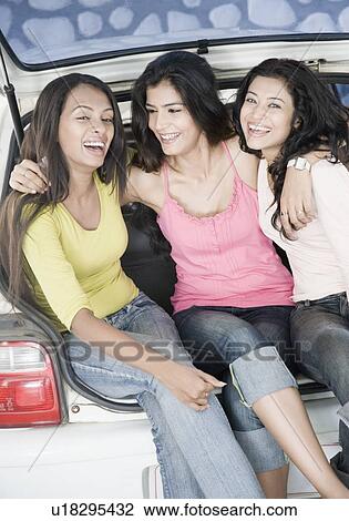 Stock Image - Three young women sitting in a car trunk and laughing. Fotosearch