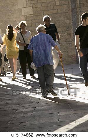 Stock Image - Old man with cane walks down crowded old section of Barcelona, Spain, in Barri Gotic area which is also known as the Gothic Quarter. Fotosearch