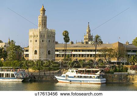 Stock Photograph - View of the tour boats and octagonal tower of Torre del Oro makes golden reflection on Canal de Alfonso of Rio Guadalquivir River, Sevilla Spain. Fotosearch