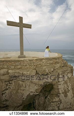 Stock Photograph - Woman stands near Cross and overlooks Atlantic Ocean at Cruz do Remedios, near Peniche, west coast of Portugal. Fotosearch