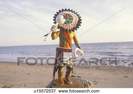 Stock Photo - Hopi Kachina doll holding objects on beach with ocean in the background. Fotosearch