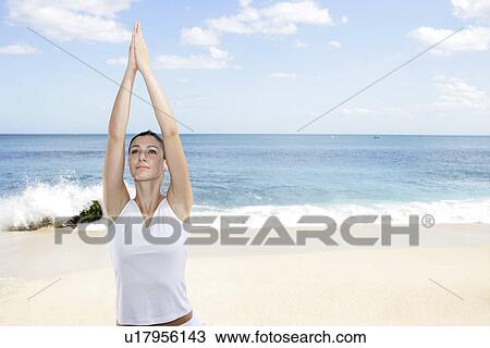 Stock Image - A woman exercising on a beach with hands raised.. Fotosearch
