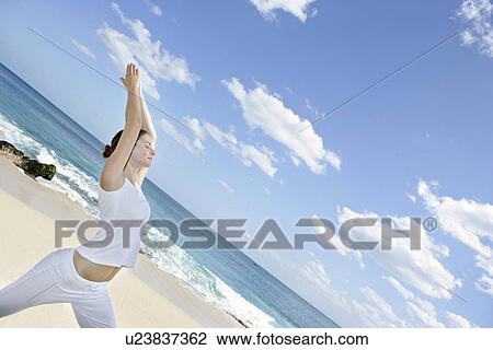 Stock Image - A woman exercising on a beach with hands raised.. Fotosearch
