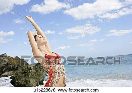 Stock Photo - A young woman standing on a beach with hand raised.. Fotosearch