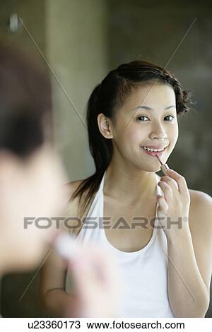 Stock Image - Mirror image of a young woman applying a lipstick.. Fotosearch