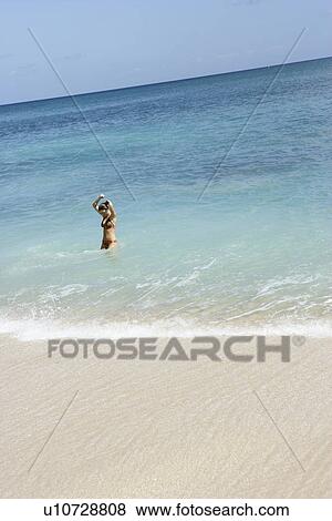 Stock Photo - View of a woman standing in sea water with hands raised.. Fotosearch