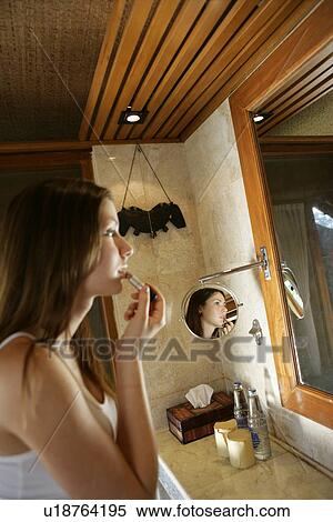 Stock Photography - Young woman applying lipstick facing a mirror.. Fotosearch