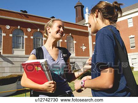 Stock Image - View of two young women conversing, holding their books.. Fotosearch