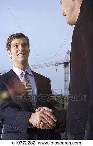 Stock Image - A Pair of Businessman Shaking Hands, Waist Up, Low Angle View, Lens Flare. Fotosearch