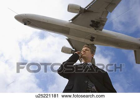 Stock Image - A Telephone Calling Businessman with a Airplane in the Background, Low Angle View, Waist Up. Fotosearch