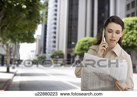 Stock Image - A Young Adult Businesswoman Looking Newspaper While Talking on the Phone, Waist Up, Differential Focus, Front View. Fotosearch