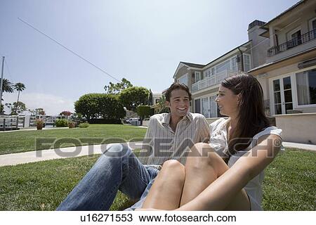 Stock Image - Young adult couple sitting on grass, Low Angle View. Fotosearch