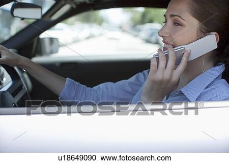 Stock Image - Young adult woman talking on mobile phone in car, Side View, Differential Focus. Fotosearch