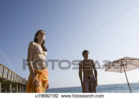 Stock Image - Young adult couple looking away on beach, Low Angle View, Copy Space. Fotosearch