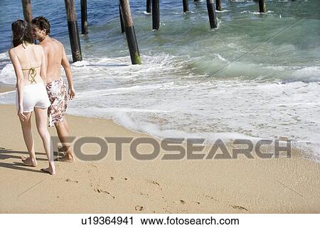 Stock Image - Young adult couple walking on beach, Rear View. Fotosearch