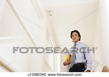 Stock Photo - Low angle view of a male architect holding rolled up blueprints and a hardhat. Fotosearch