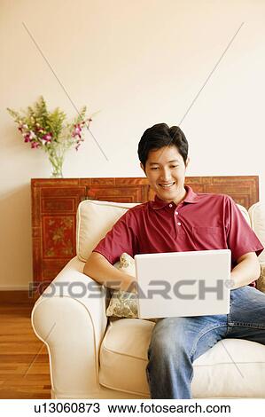 Young man sitting on a couch and using a laptop View Large Photo Image Stock Image - Young man sitting on a couch and using a laptop. Fotosearch