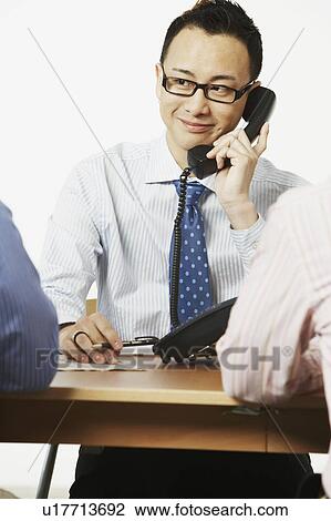 Close-up of a businessman talking on the telephone with two businessmen sitting in front of him View Large Photo Image Stock Image - Close-up of a businessman talking on the telephone with two businessmen sitting in front of him. Fotosearch
