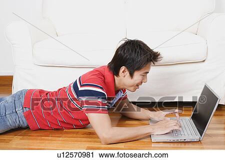 Stock Image - Side profile of a mid adult man lying on the floor and using a laptop. Fotosearch