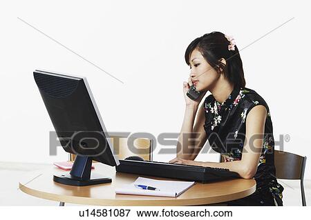 Young woman sitting in front of a computer talking on the telephone View Large Photo Image Stock Photo - Young woman sitting in front of a computer talking on the telephone. Fotosearch