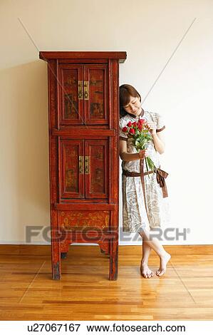 Stock Photo - Close-up of a young woman standing near a cabinet and holding a bouquet of red roses. Fotosearch