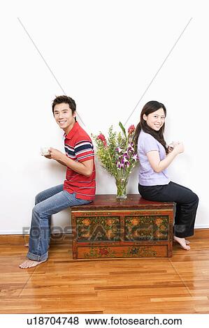 Stock Photo - Portrait of a mid adult man and a young woman sitting on a trunk and holding cups of tea. Fotosearch