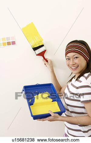 Stock Photo - Portrait of a young woman painting a wall with a paintbrush and smiling. Fotosearch