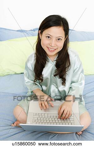 Stock Image - Portrait of a young woman using a laptop on the bed and smiling. Fotosearch