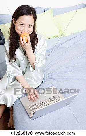 Stock Image - Portrait of a young woman using a laptop on the bed and holding an orange. Fotosearch