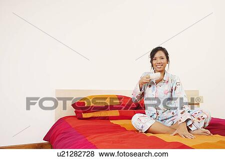 Stock Photo - Young woman sitting on the bed and holding a cup of tea. Fotosearch
