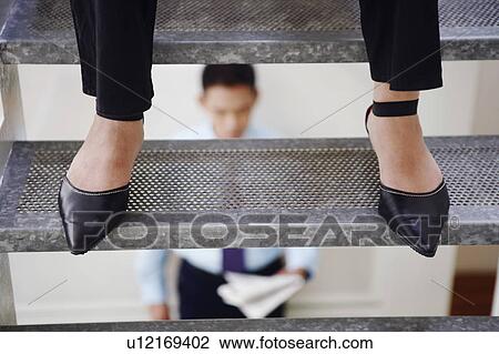 Stock Image - Low section view of a person standing on a staircase and a young man reading a newspaper in background. Fotosearch