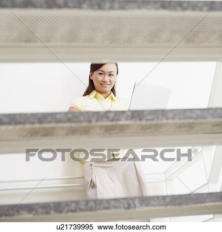 Stock Photography - Portrait of a young woman leaning against a wall and using a laptop. Fotosearch