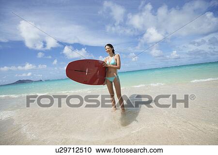 Stock Image - A young woman holding surfboard on beach. Fotosearch