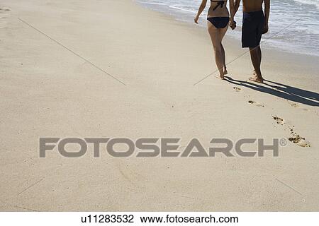 Stock Image - Low section of a couple walking at beach. Fotosearch
