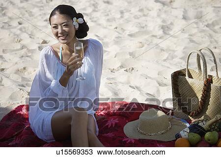 Stock Image - A young woman holding wineglass and sitting at beach. Fotosearch