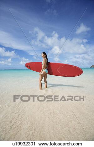 Stock Image - Profile of young woman holding surfboard at beach. Fotosearch