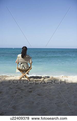 Stock Photo - Rear view of a woman sitting on chair at beach. Fotosearch