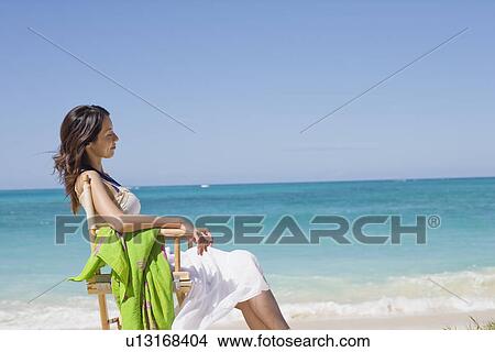 Side view of a young woman sitting on chair at beach View Large Photo Image Picture - Side view of a young woman sitting on chair at beach. Fotosearch