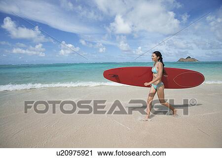 Stock Image - A young woman walking on beach with surfboard. Fotosearch