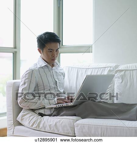 Young man sitting on a couch using a laptop View Large Photo Image Stock Image - Young man sitting on a couch using a laptop. Fotosearch