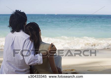 Stock Photography - Rear view of a young couple sitting on beach. Fotosearch