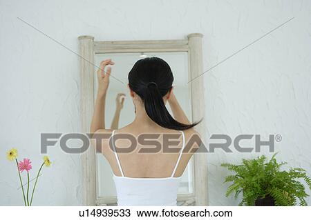 Stock Image - Rear view of a young woman standing in front of mirror. Fotosearch