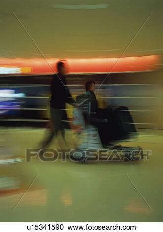 Stock Image - Travelers walking with luggage cart at airport, blurred motion. Fotosearch