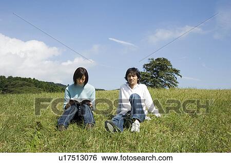 Stock Photograph - Young couple sitting on field, woman reading book. Fotosearch