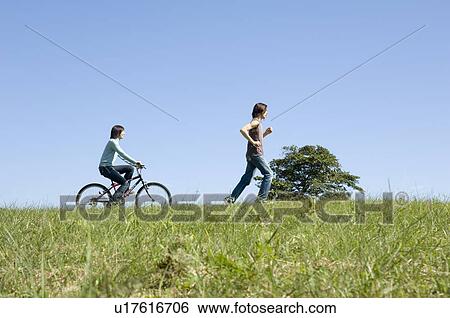 Young woman riding bicycle behind young man running View Large Photo Image Stock Photograph - Young woman riding bicycle behind young man running. Fotosearch